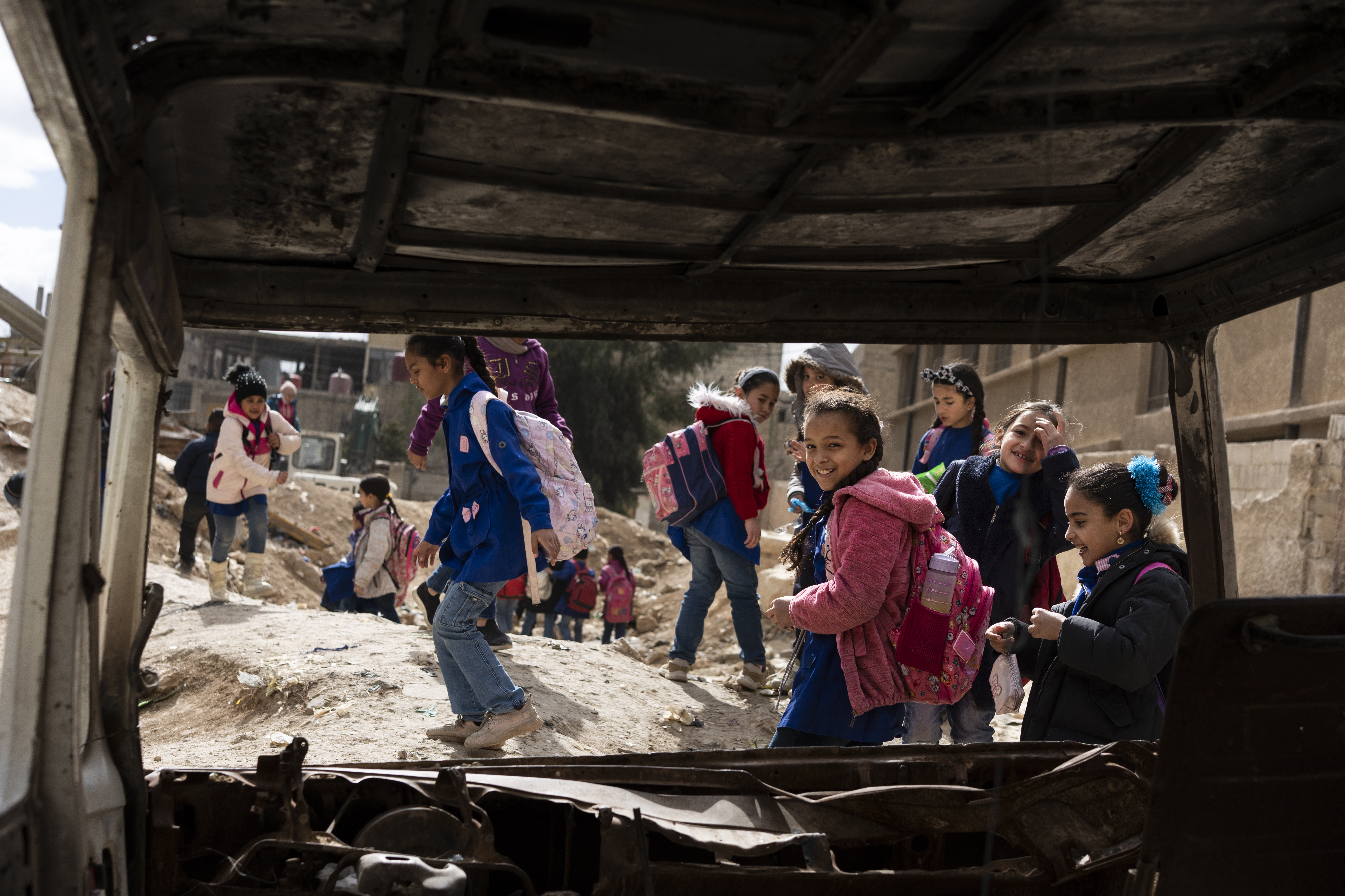 Schoolgirls in a street devastated by bombing, photographed through the wreckage of a burnt-out car.