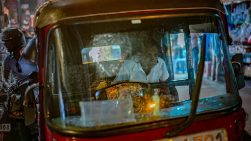 A toddler in red stares wide-eyed from a passenger's lap inside a black tuk-tuk, the city's night lights glowing through the open side.