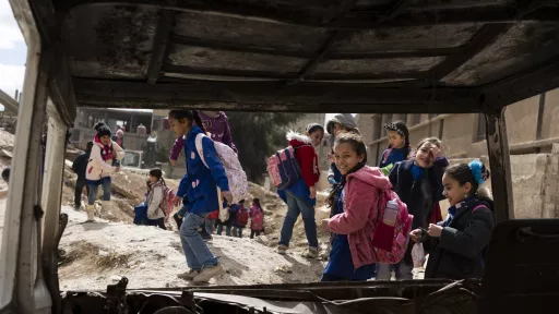 Schoolgirls in a street devastated by bombing, photographed through the wreckage of a burnt-out car.