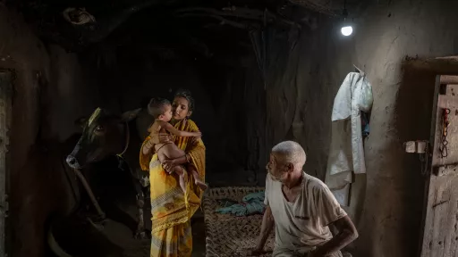 A woman holds a small child inside a dim mud-walled room with a cow, while an old man sits on a rope cot.