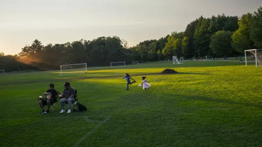 Family picnic in a park at sunset, people seated at a picnic table and in folding chairs near soccer goals, coolers and food spread on a table.