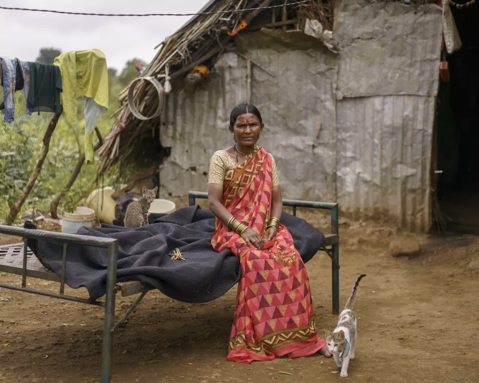 Rural woman wearing a red patterned sari seated on a simple metal bed outside a small cement house
