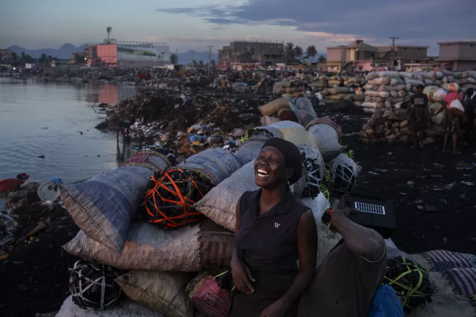 Woman in dark vest laughing joyfully while sitting among large wrapped bundles at waterfront during dusk