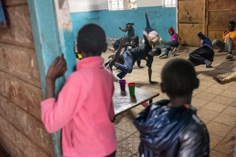 Indoor gymnasium with teal walls where youth in white clothing perform synchronized backflips