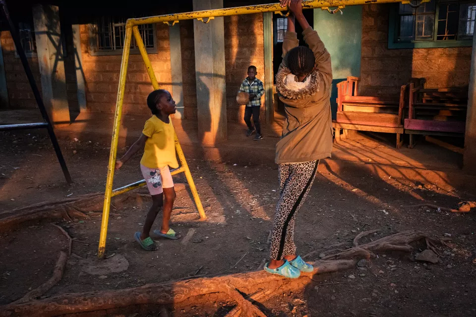 Woman performing pull-ups on a yellow chin-up bar in a settlement courtyard during golden hour