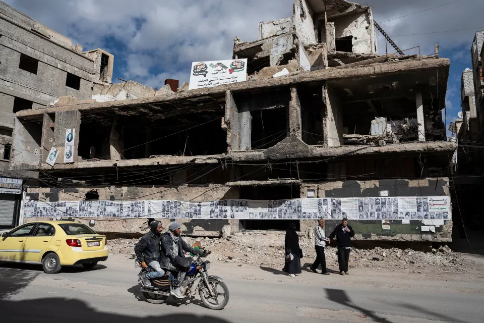 A devastated street with a motorcycle and a car, passersby. On a wall, numerous portraits of missing martyrs.