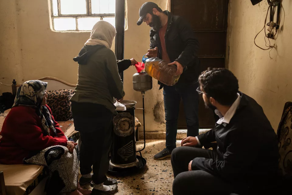 In a very poor interior, two volunteers help an elderly couple  fill their old kerosene stove.
