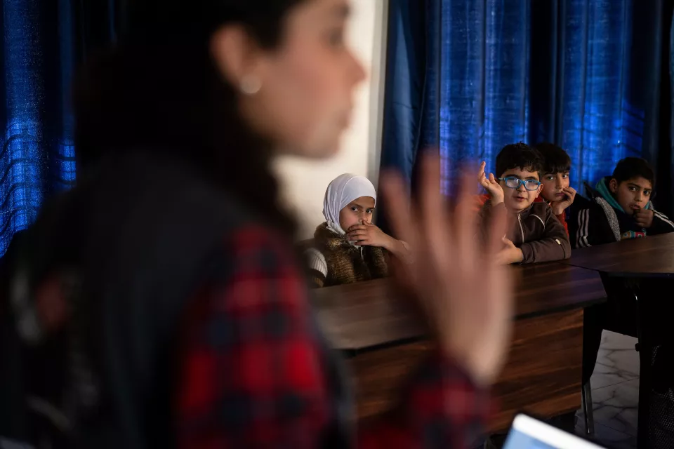 Four children in a classroom listen to a volunteer in the foreground who introduces the film screening.
