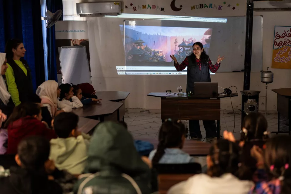 A volunteer, arms outstretched in front of a screen, presents the film for the session to children seated in a U-shape around the projector. 