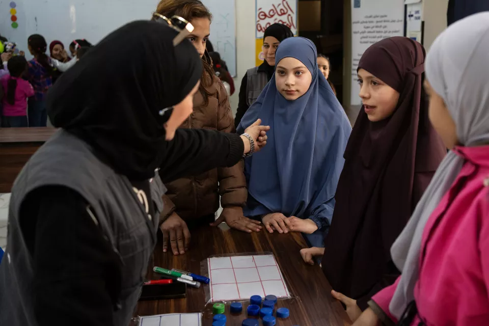 Young girls beautifully veiled in blue, burgundy, and pink stand around a table listening to instructions from a volunteer.