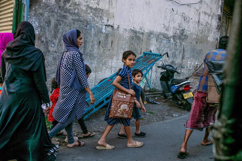 A woman in a hijab walks with children through a back alley, a young girl glancing back at the camera beside a leaning blue rack and parked scooter.