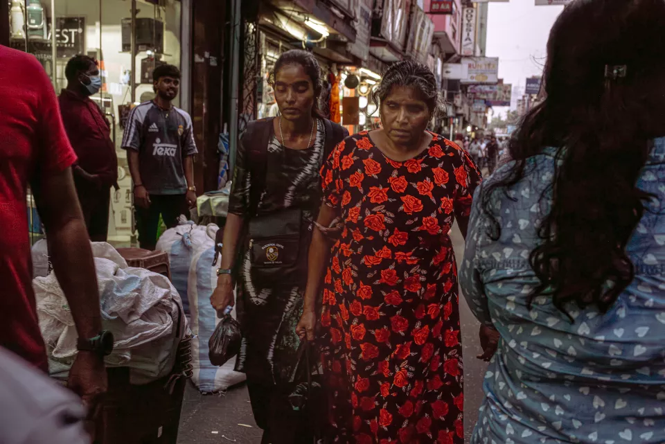 Two women navigate a narrow, bustling market street in Colombo, the bold red roses of one woman's dress vivid against the crowded backdrop.