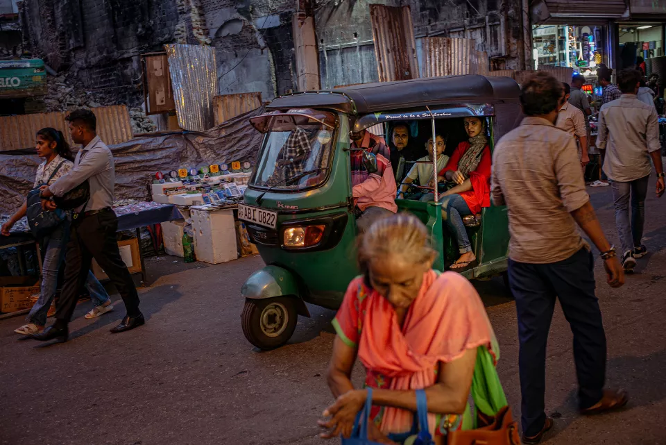 A green tuk-tuk packed with passengers edges through evening traffic, an elderly woman in pink crossing in the foreground amid the crowd.