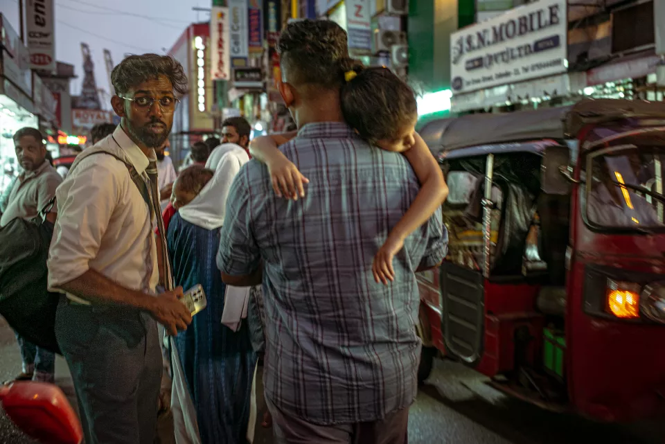 A man in a loosened tie glances sideways at the camera while another carries a sleeping child through the neon-lit chaos of a Colombo night street.