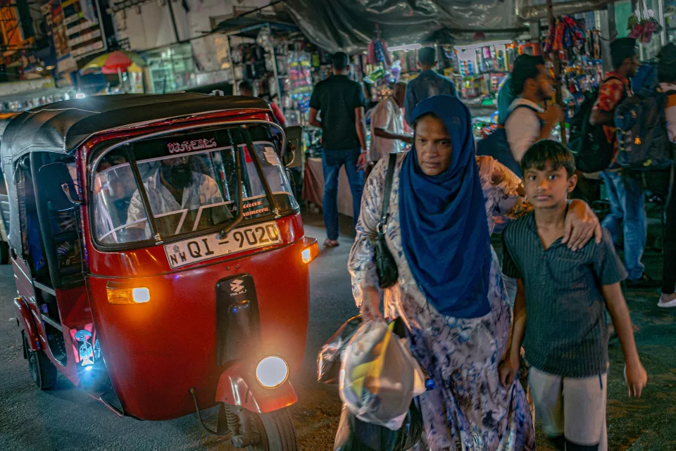 A woman in a blue hijab steers her son through a packed night market, a glowing red tuk-tuk pressing close behind them.