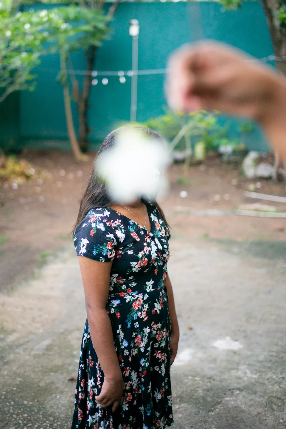 A woman in a floral dress stands in a garden, her face obscured by a flower held close to the lens &mdash; a playful, poetic composition echoing the earlier garden portrait.