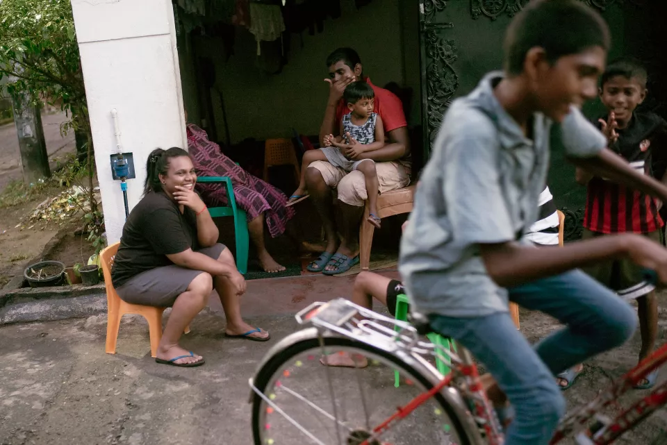 A family relaxes on their doorstep, smiling as boys cycle past on a quiet Colombo street &mdash; an unhurried moment of everyday neighborhood life.