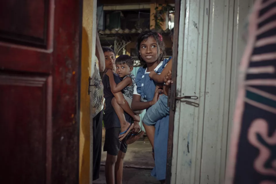 Three children peer curiously through a half-open doorway, one girl leaning boldly on the frame with a wide, eager smile.