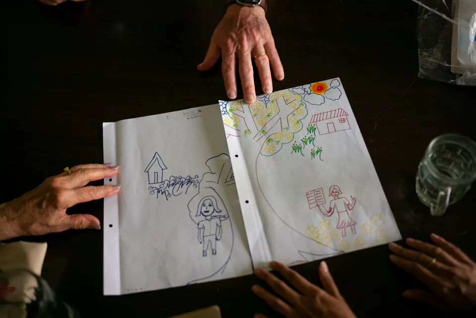 Two adults hold open a notebook filled with children's drawings &mdash; figures, houses, and trees &mdash; suggesting a participatory or educational workshop.