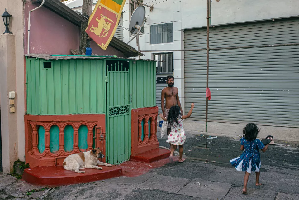 A shirtless man watches two young girls play in front of a vivid green and red street shrine, a Sri Lankan flag flying overhead.