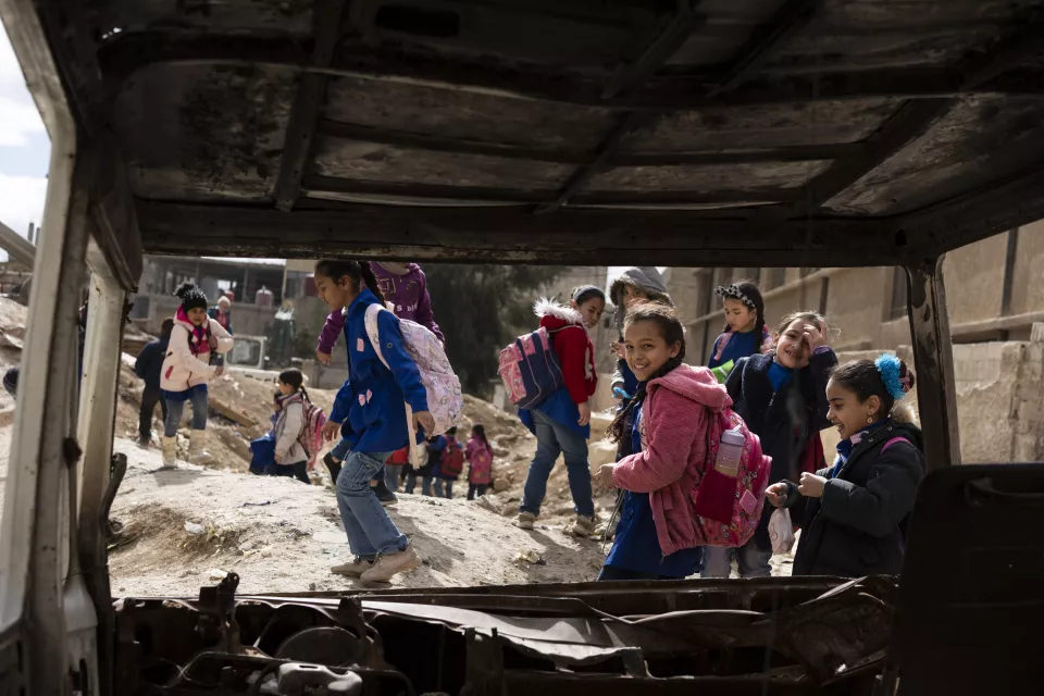 Schoolgirls in a street devastated by bombing, photographed through the wreckage of a burnt-out car.
