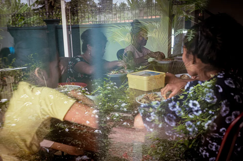 Women share a meal around a table, seen through a window layered with reflections of lush garden foliage, blending interior and exterior.