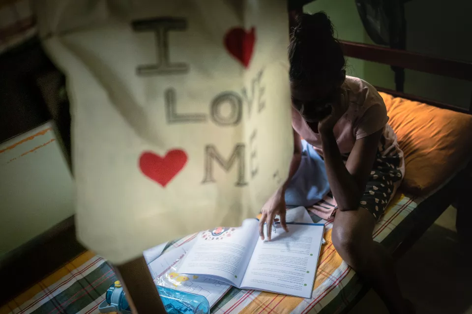 A young girl studies intently at a bedside table in a dimly lit room, an "I Love Me" tote bag hanging in the foreground.