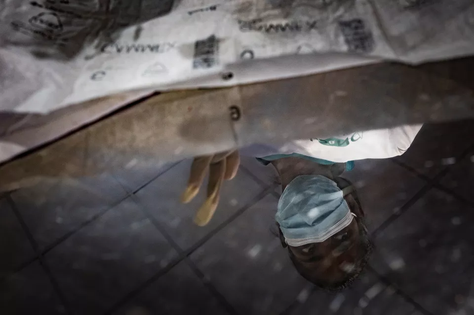 A disorienting close-up reflection on a wet tiled floor, showing a gloved hand and a surgical mask dangling beneath crumpled packaging.