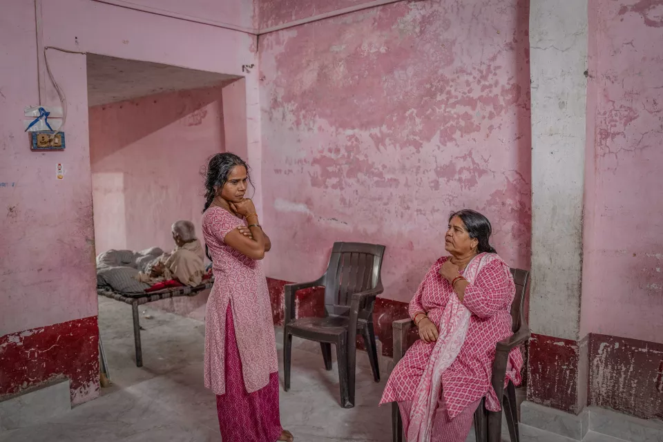 Two women in pink clothing talk in a pink room with peeling walls; an elderly man rests on a cot behind them.
