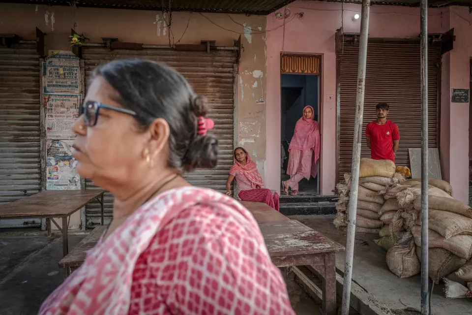 A woman in glasses and a pink-checked top walks past in the foreground, while two women and a man in red watch from a pink shopfront.