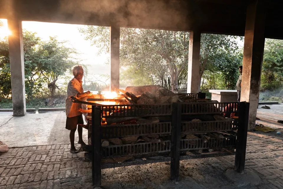 An elderly man tends a wood funeral pyre burning inside an open-air cremation shelter at sunrise.