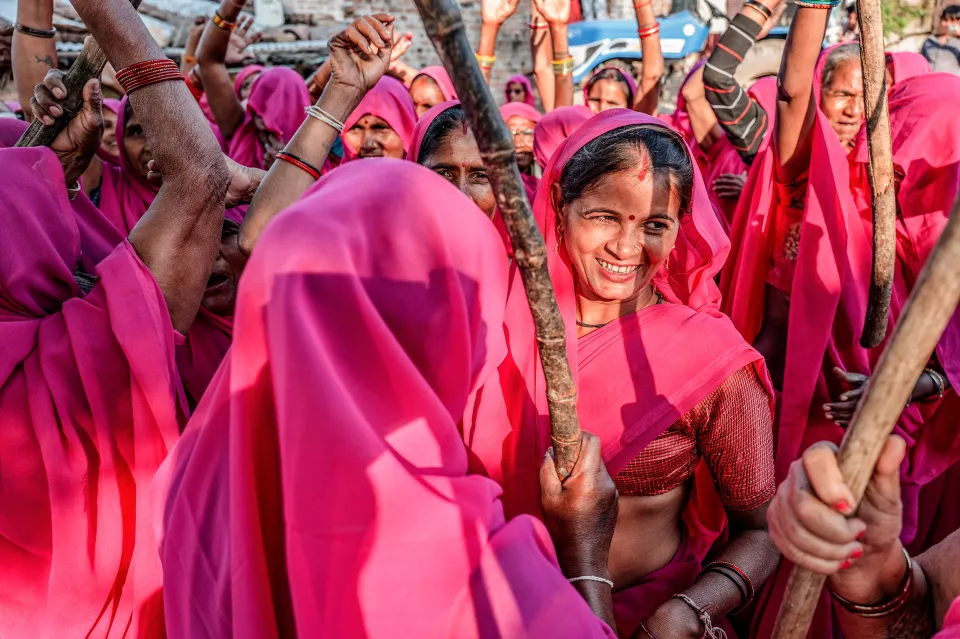 Smiling woman in a pink sari raises a wooden stick amid a crowd of women in pink saris with raised arms.