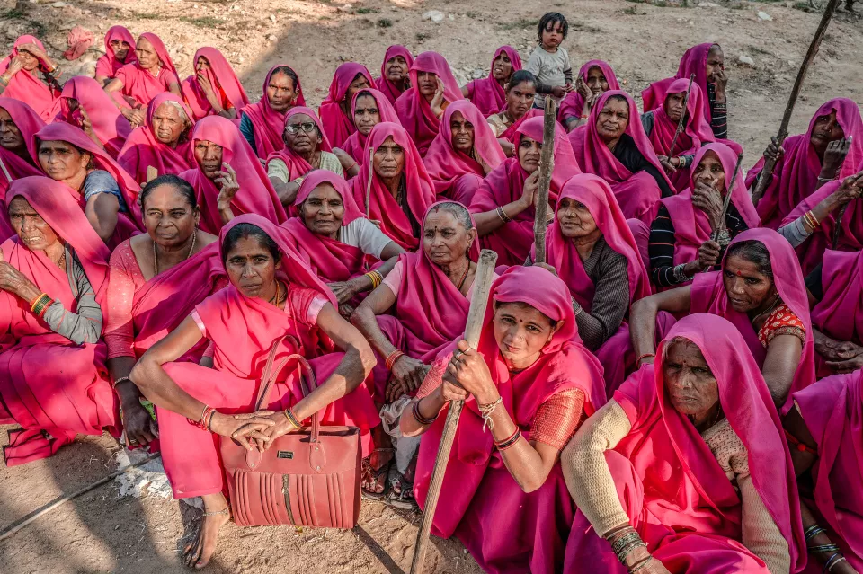 A large group of women in pink saris sit on dry ground, some holding wooden sticks, a small child among them.