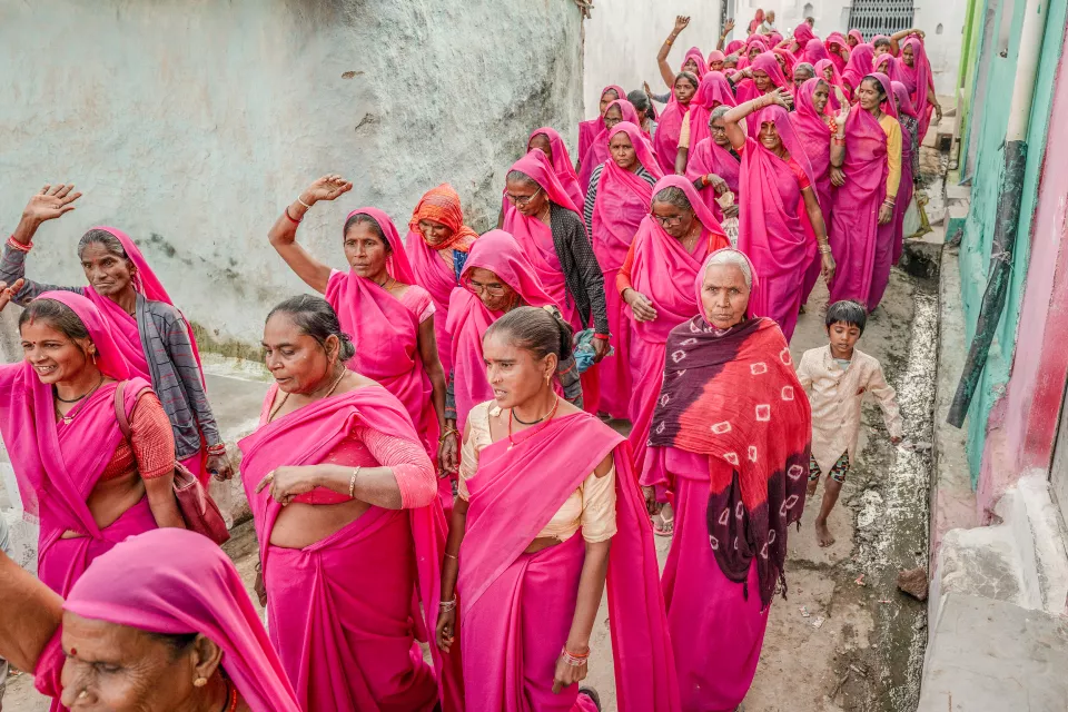 Women in pink saris march in a narrow lane, some raising their hands, a young boy walking beside them.