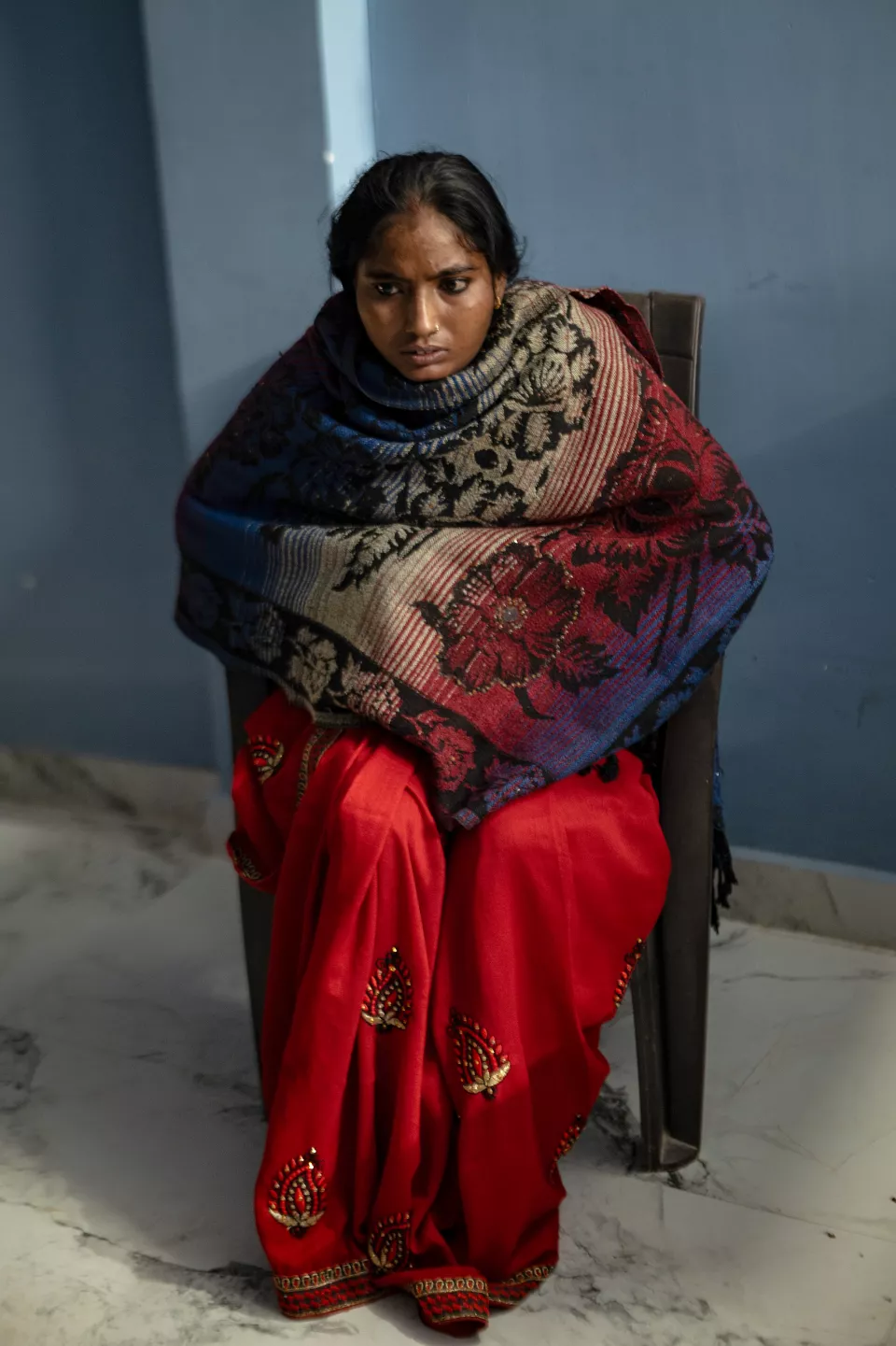 A young woman in a red sari, wrapped in a patterned shawl, sits on a chair against a blue wall, looking worried.