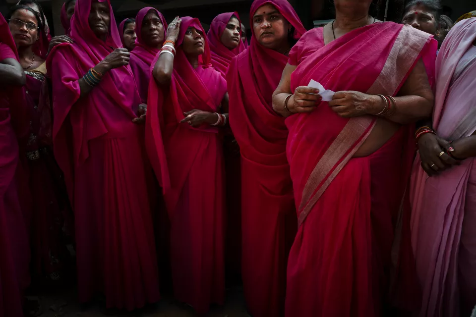 Women in pink saris stand close together in shadow; one in the foreground holds a small piece of paper.