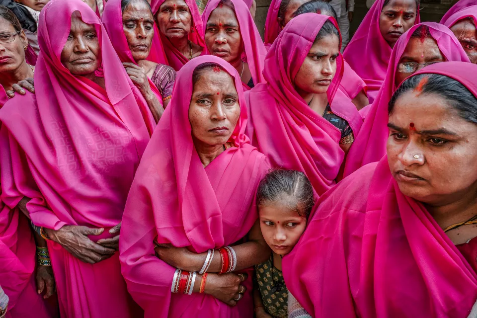 Close-up of women in pink saris with serious faces, a young girl leaning against one of them.
