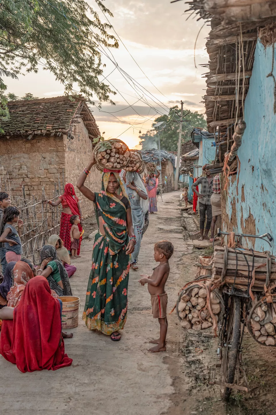 A woman balances a basket of firewood on her head in a village lane at dusk; a small boy and seated women look on.