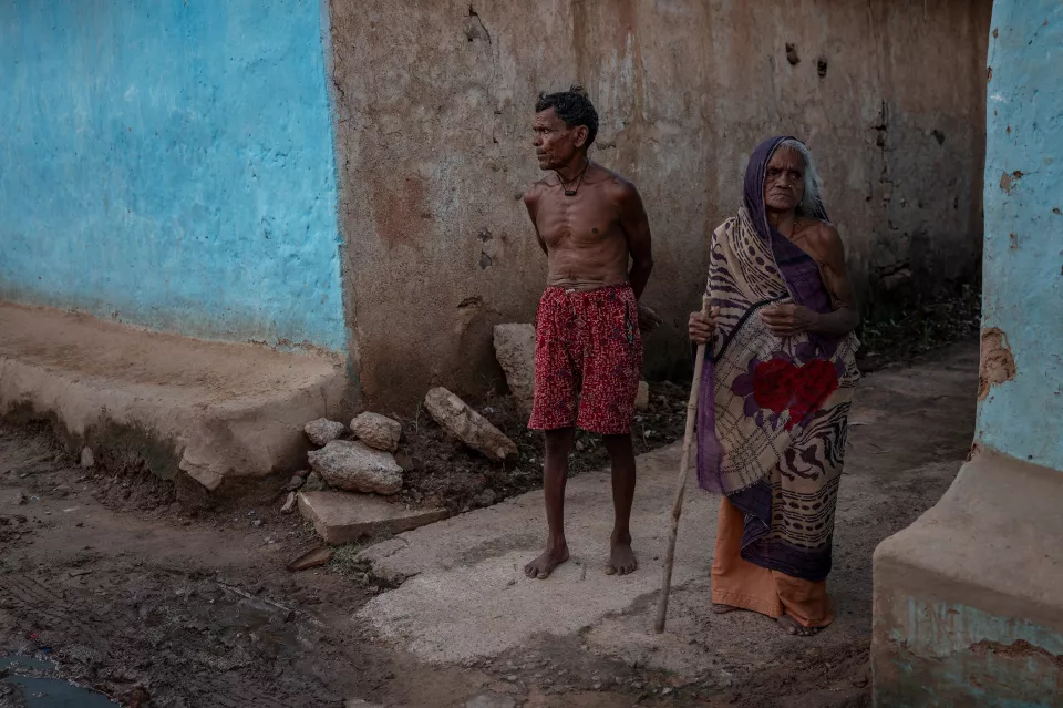 An elderly man and woman stand side by side beside blue and brown mud walls; she leans on a wooden stick.