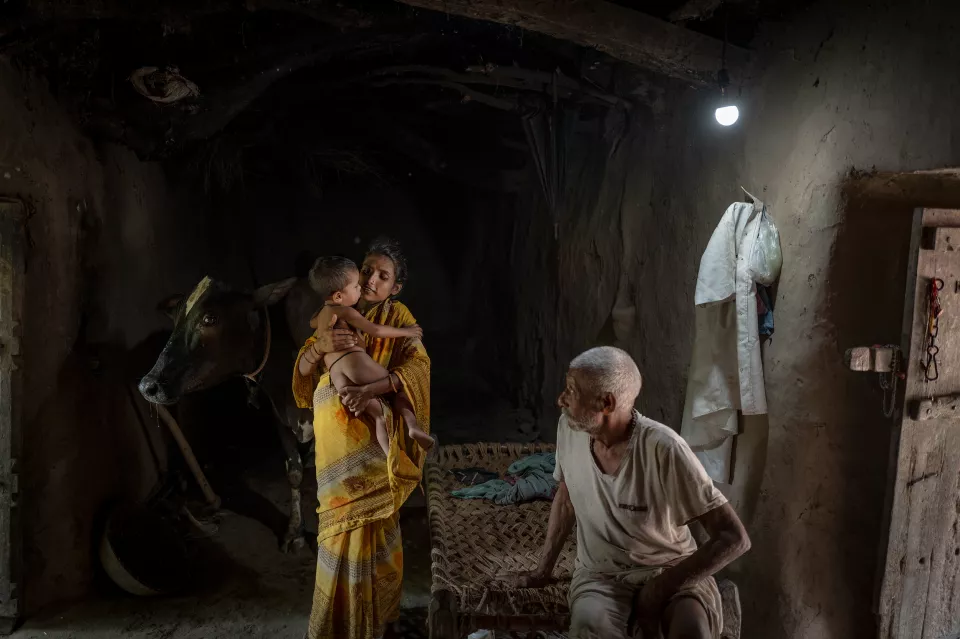 A woman holds a small child inside a dim mud-walled room with a cow, while an old man sits on a rope cot.