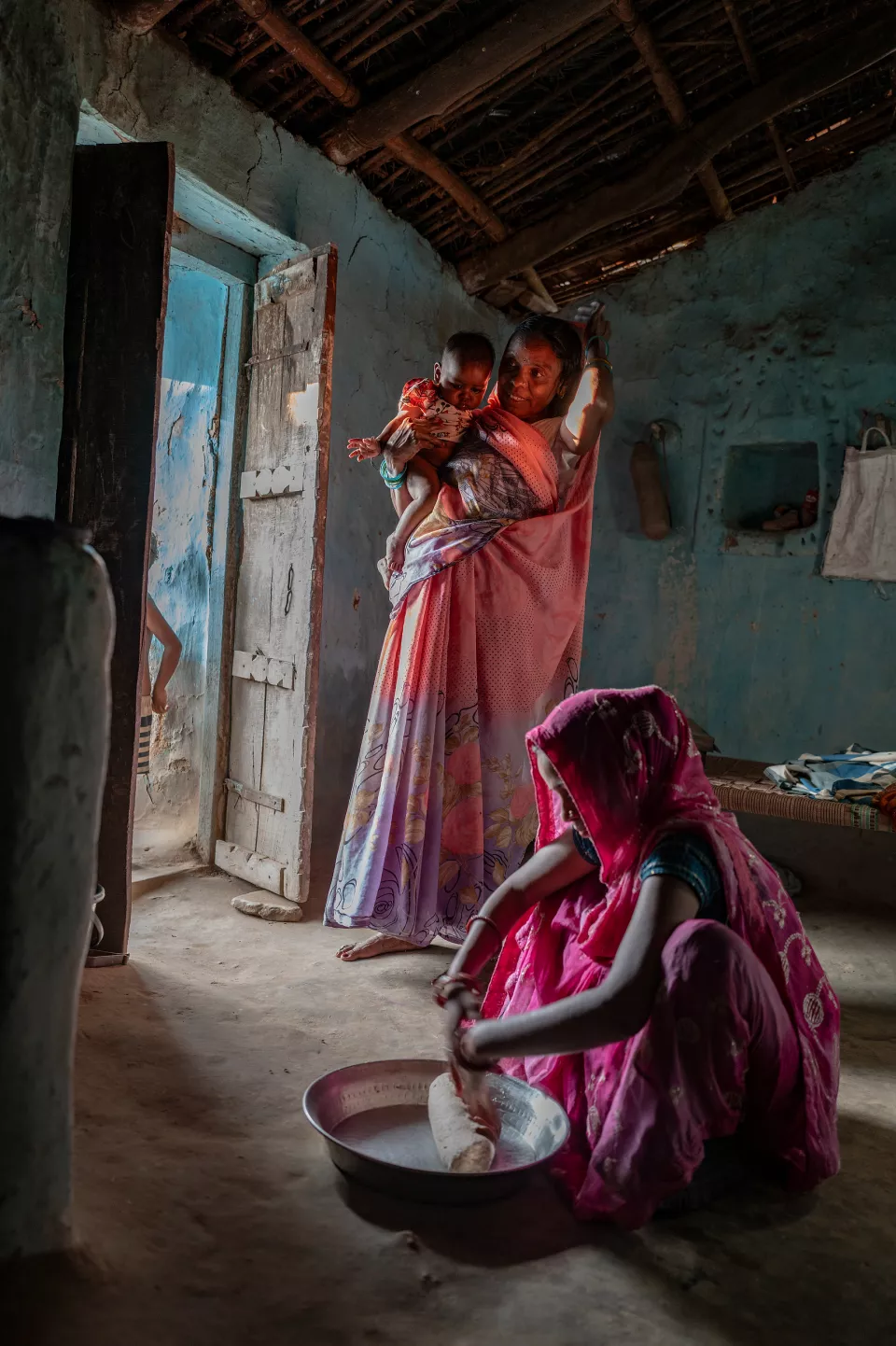 A woman in a pink shawl kneels over a metal basin kneading dough as another woman holding a baby watches from behind.