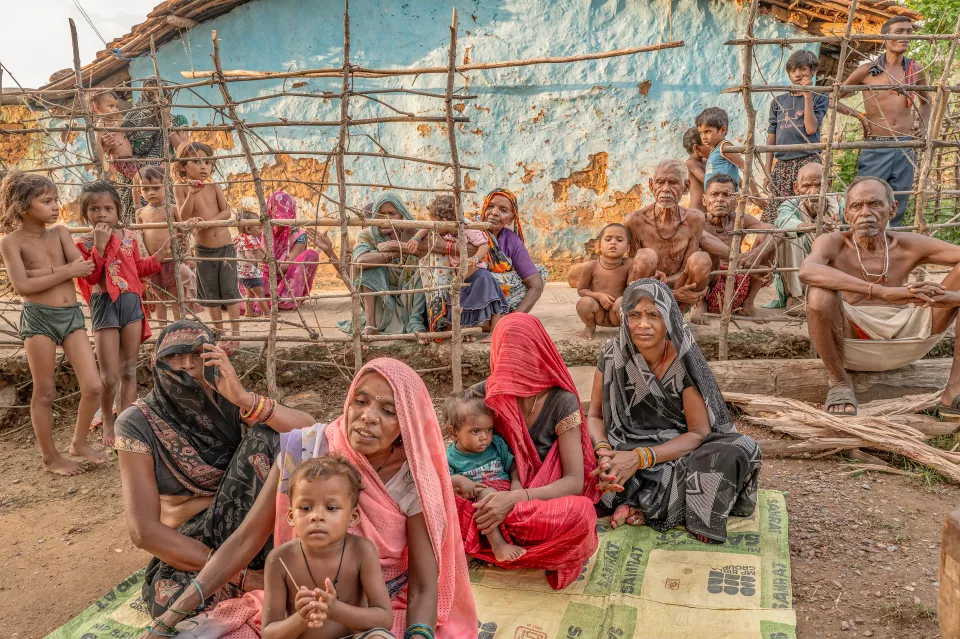 Villagers, including many children and elderly people, sit and stand behind a rough wooden fence in front of a blue wall.