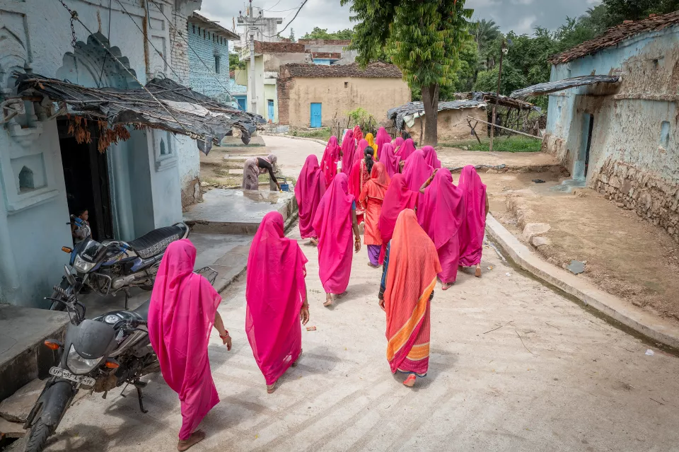 Women in bright pink saris walk away from the camera along a village lane lined with blue houses and parked motorbikes.