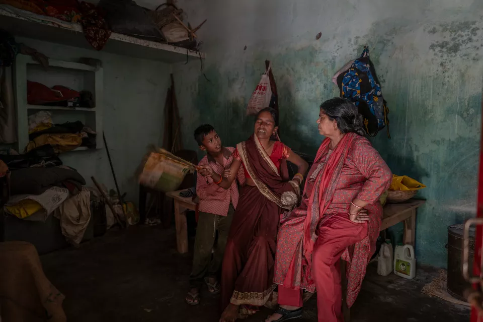 A boy holds a woven fan next to a seated woman in a brown sari as an older woman in red looks on in a green-walled room.