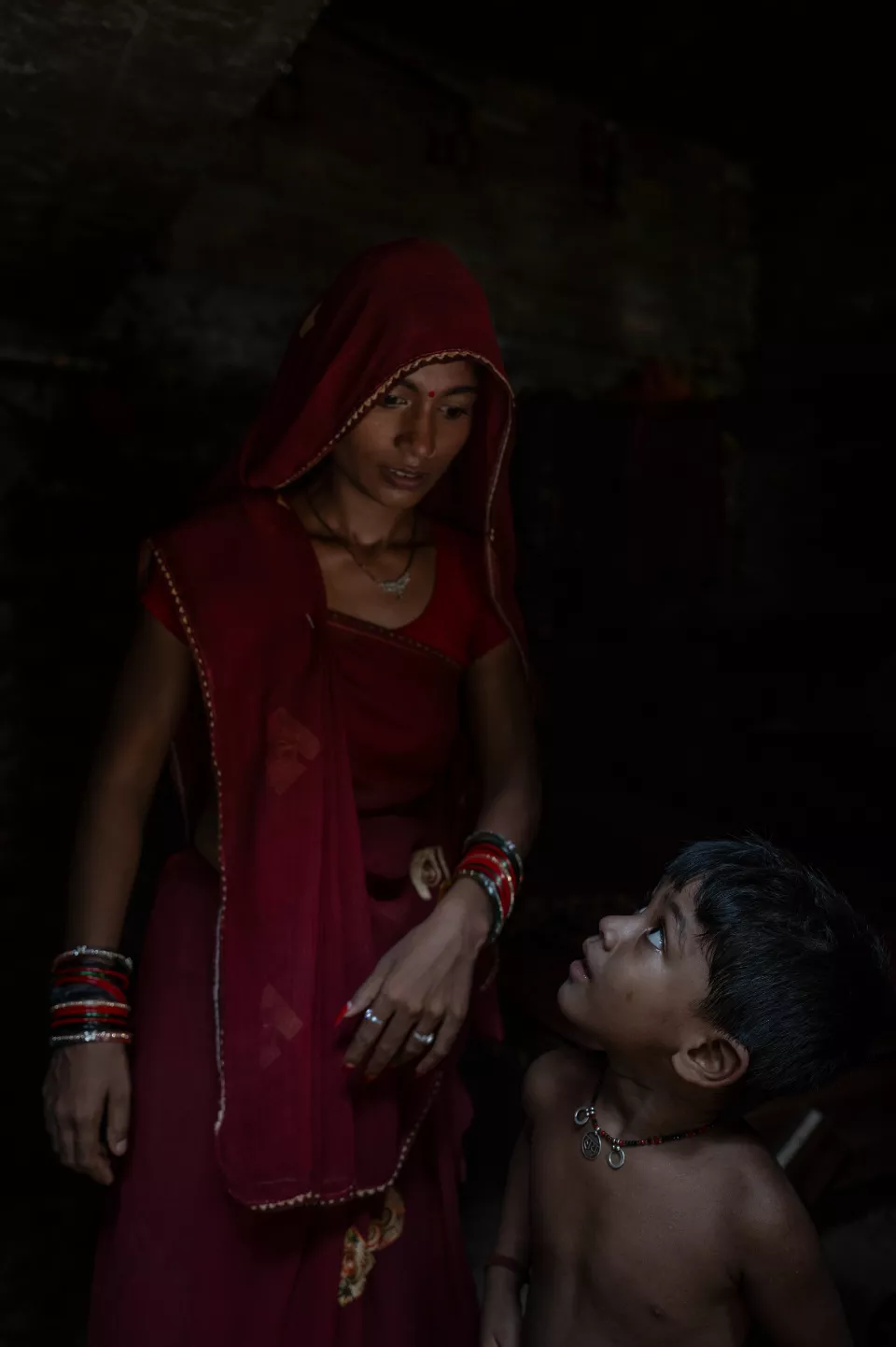 A woman in a red veil looks down at a shirtless boy who gazes up at her in a dark room.
