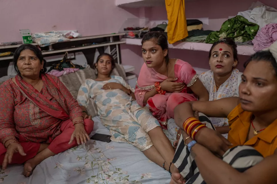 Five women sit and recline on a bed in a pink room, looking attentively in the same direction.