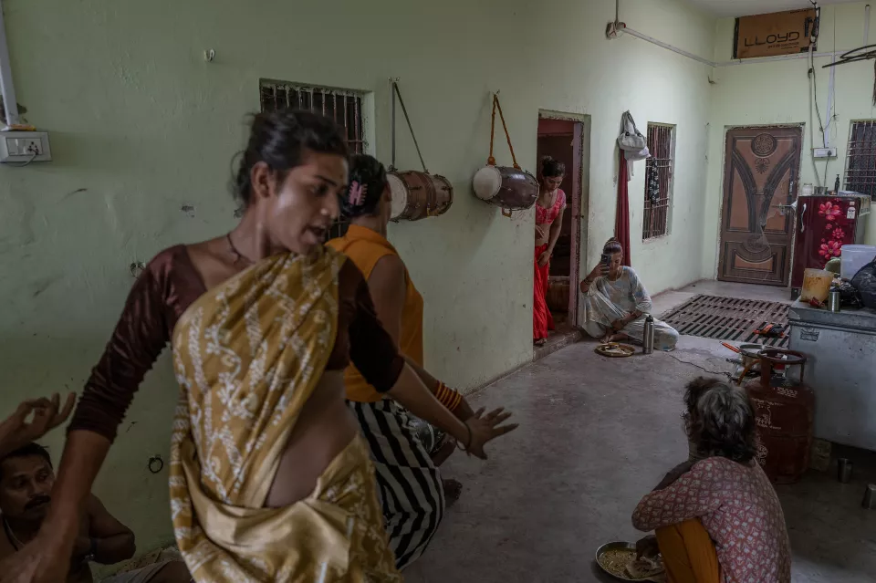 A woman in a gold-patterned sari moves energetically in a green room with hanging drums, while others eat and use a phone.