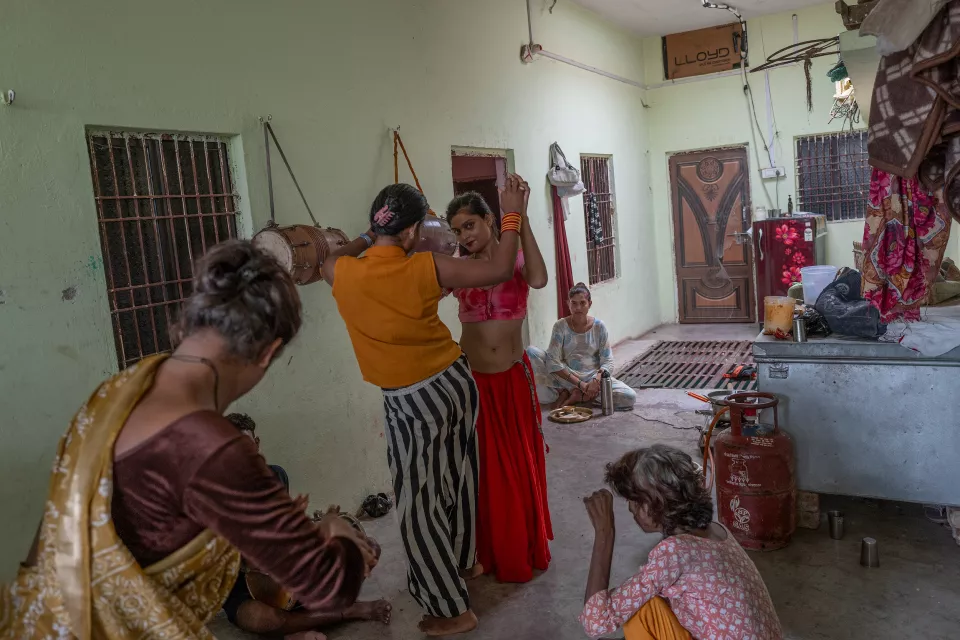 Two people dance with raised hands in a green room with hanging drums, as others watch, play a drum, and a woman sits eating on the floor.