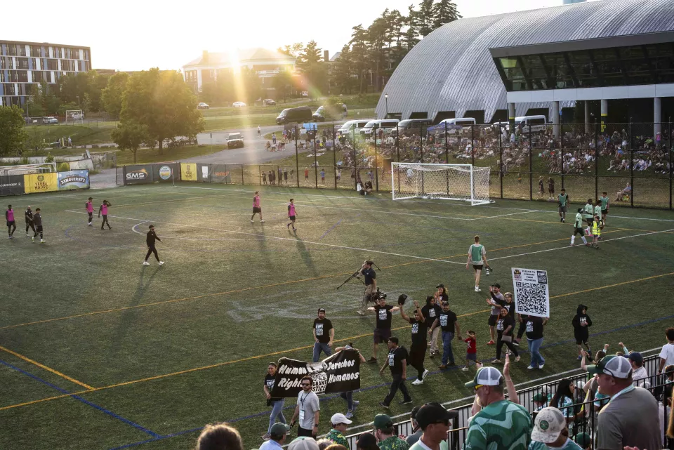 Close-up of the "Human Rights / Derechos Humanos &ndash; Migrant Justice" banner carried by volunteers across the field, crowd blurred in the stands behind.