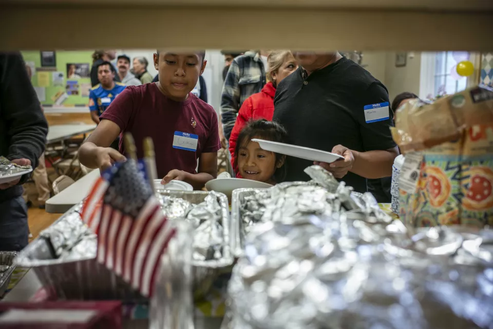 A boy and a young girl wearing name tags serve themselves from foil trays decorated with a small American flag, community hall.